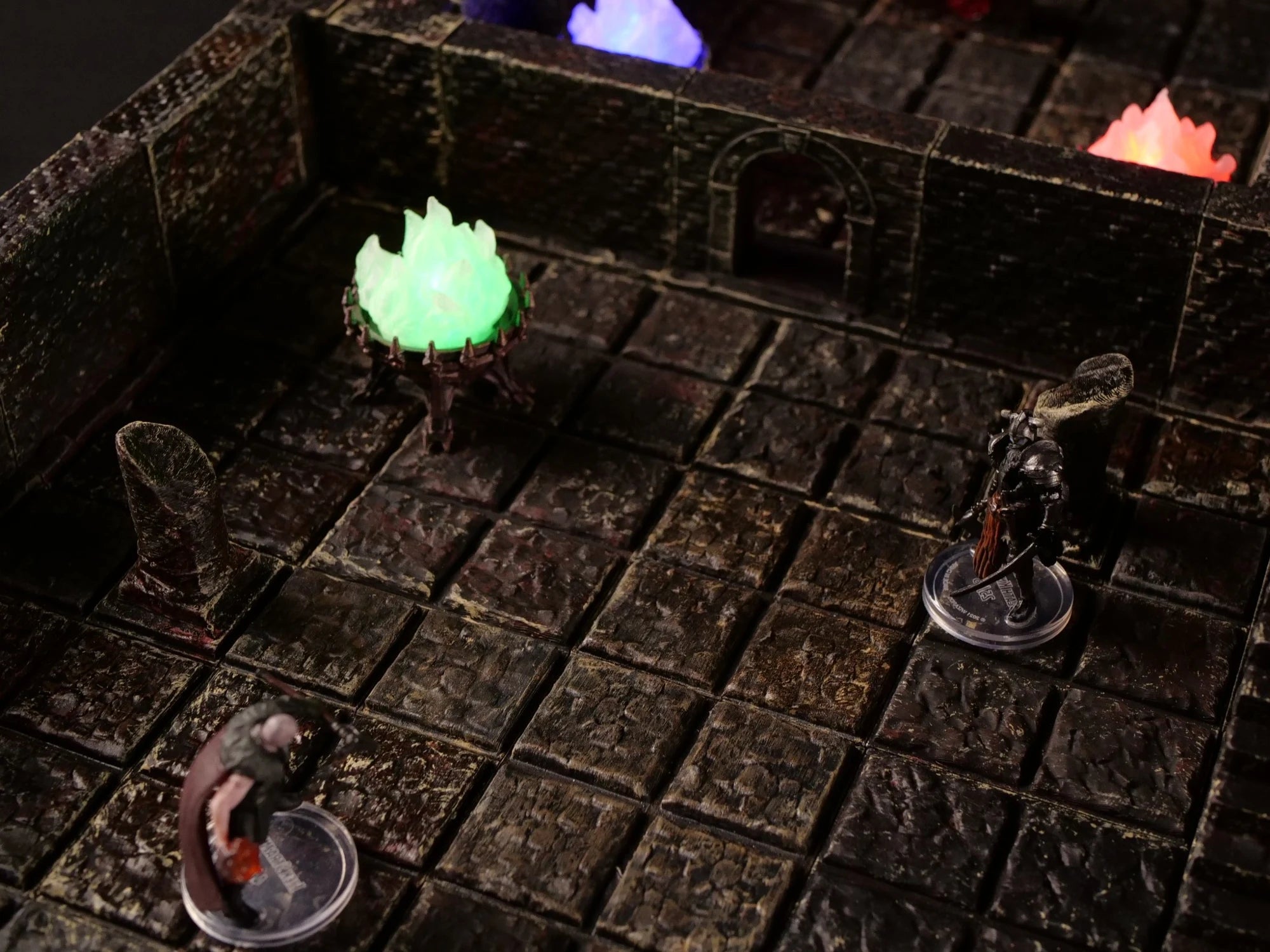 Stone dungeon terrain with miniature adventurers and glowing green and violet braziers